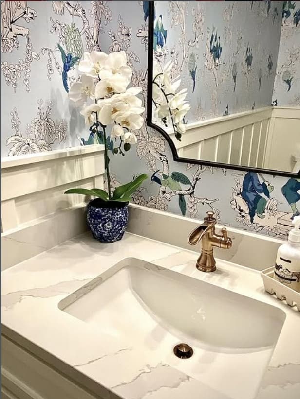 Bathroom vanity with blue botanical wallpaper, marble counter, brass faucet, and a white orchid