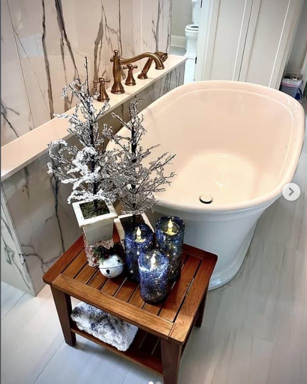 Freestanding white bathtub with marble surround, brass fixtures, a teak bench, and candlelit winter styling