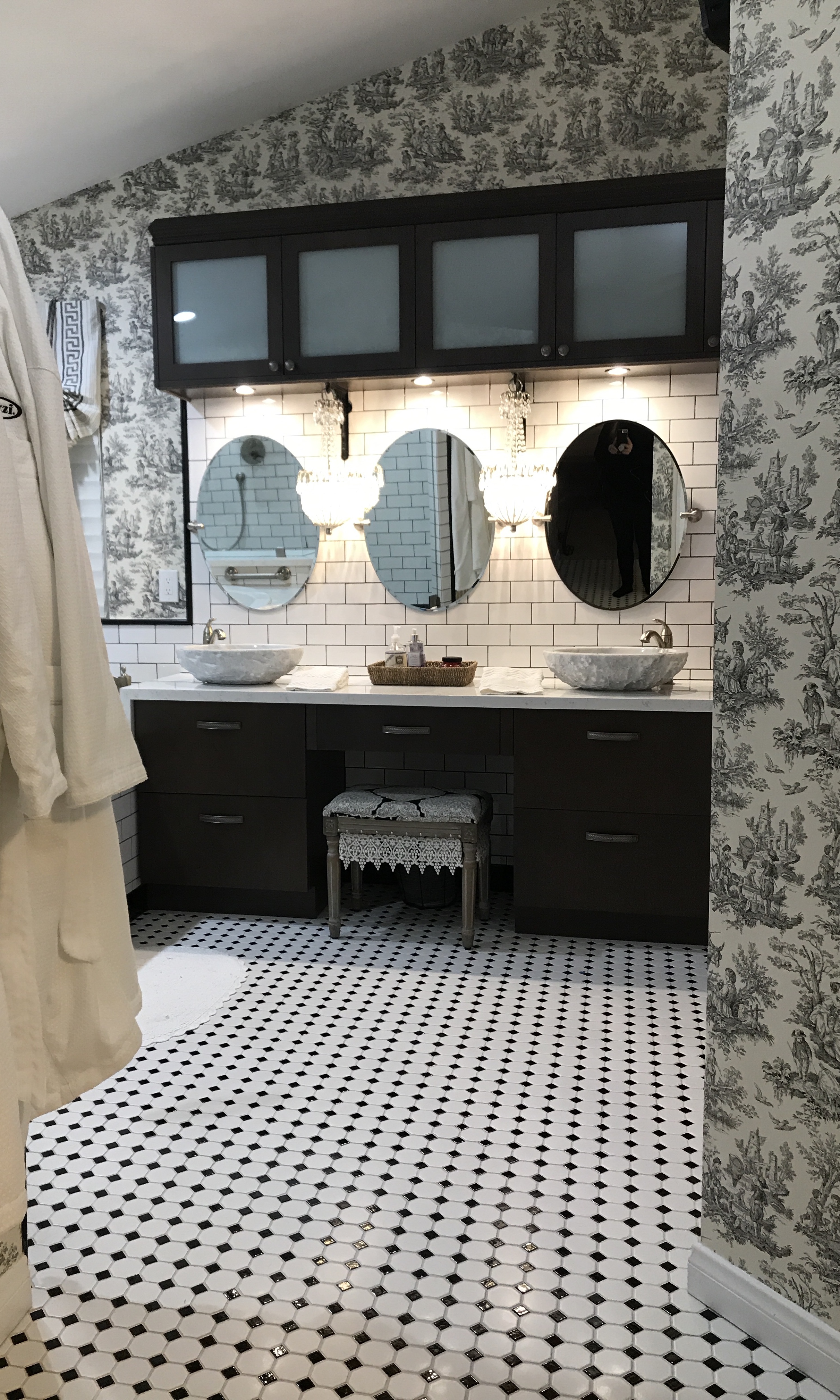 Classic black-and-white bathroom with gray toile wallpaper, dark espresso vanity with dual marble vessel sinks, three oval mirrors, crystal chandelier sconces, and a diamond mosaic tile floor