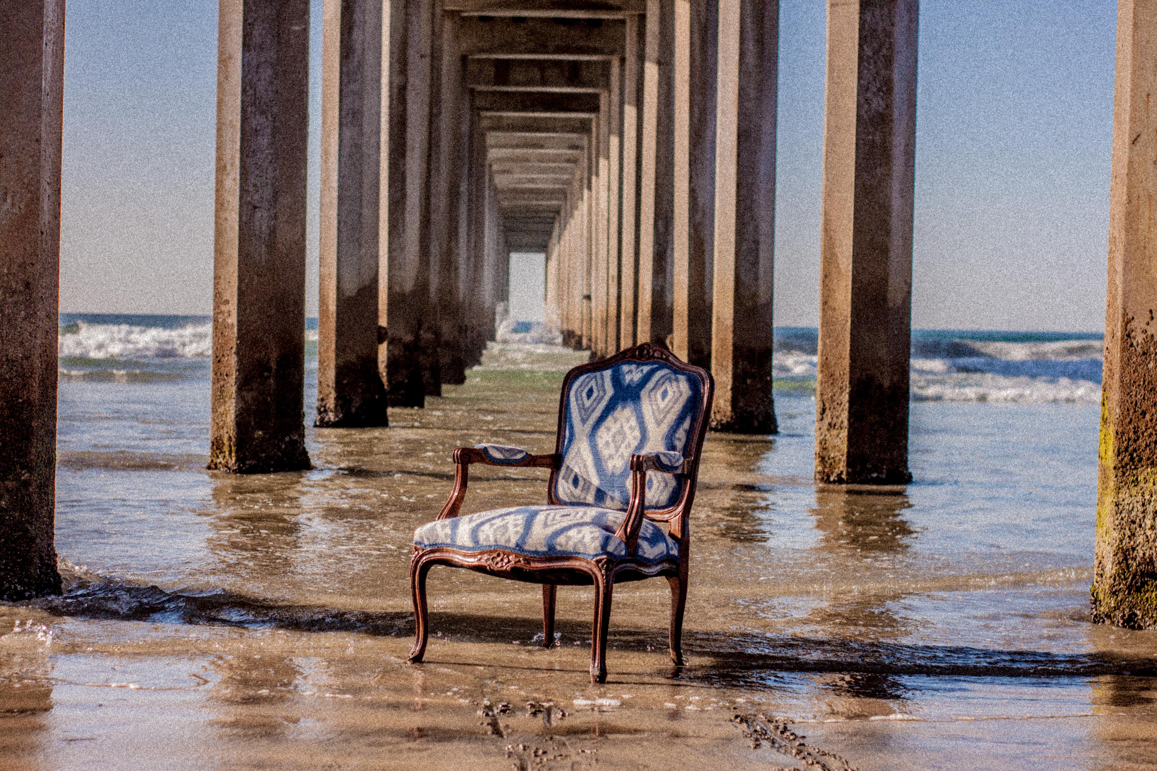 Ornate French-style wooden armchair upholstered in blue-and-white ikat fabric placed on wet sand beneath a long pier with rows of concrete pillars receding into the distance and ocean waves