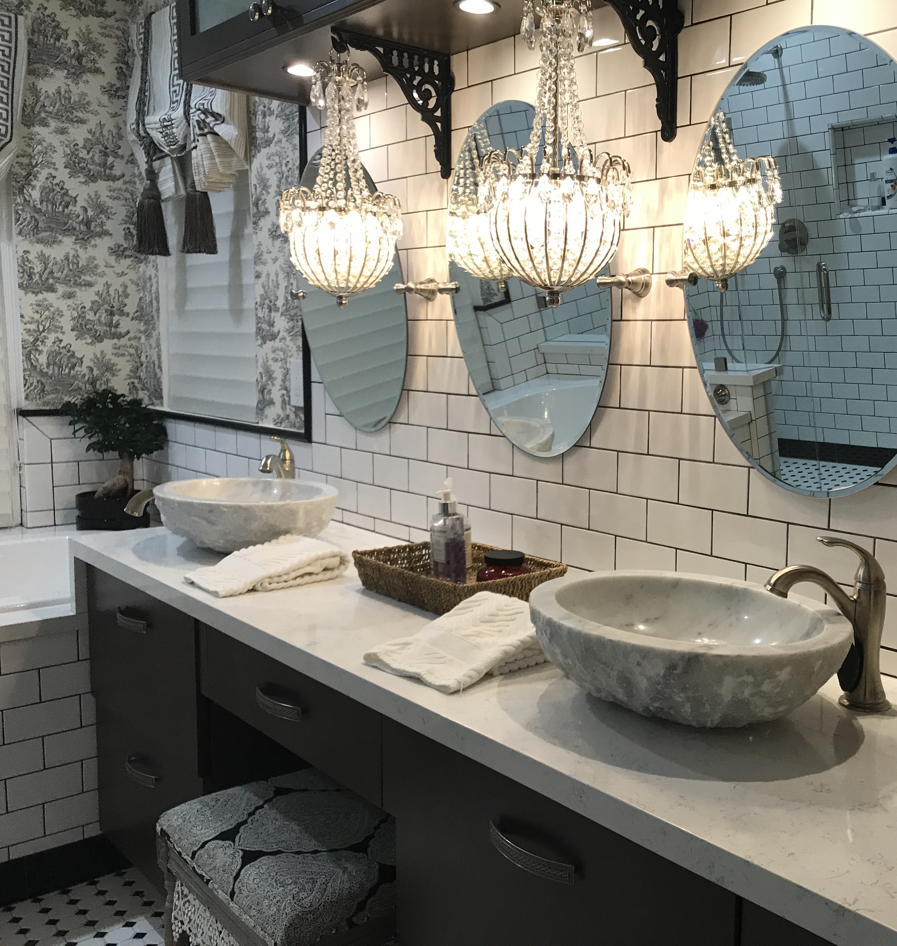 Elegant double-vanity bathroom with white subway tile, dual marble vessel sinks, crystal chandelier sconces, oval mirrors, and toile wallpaper