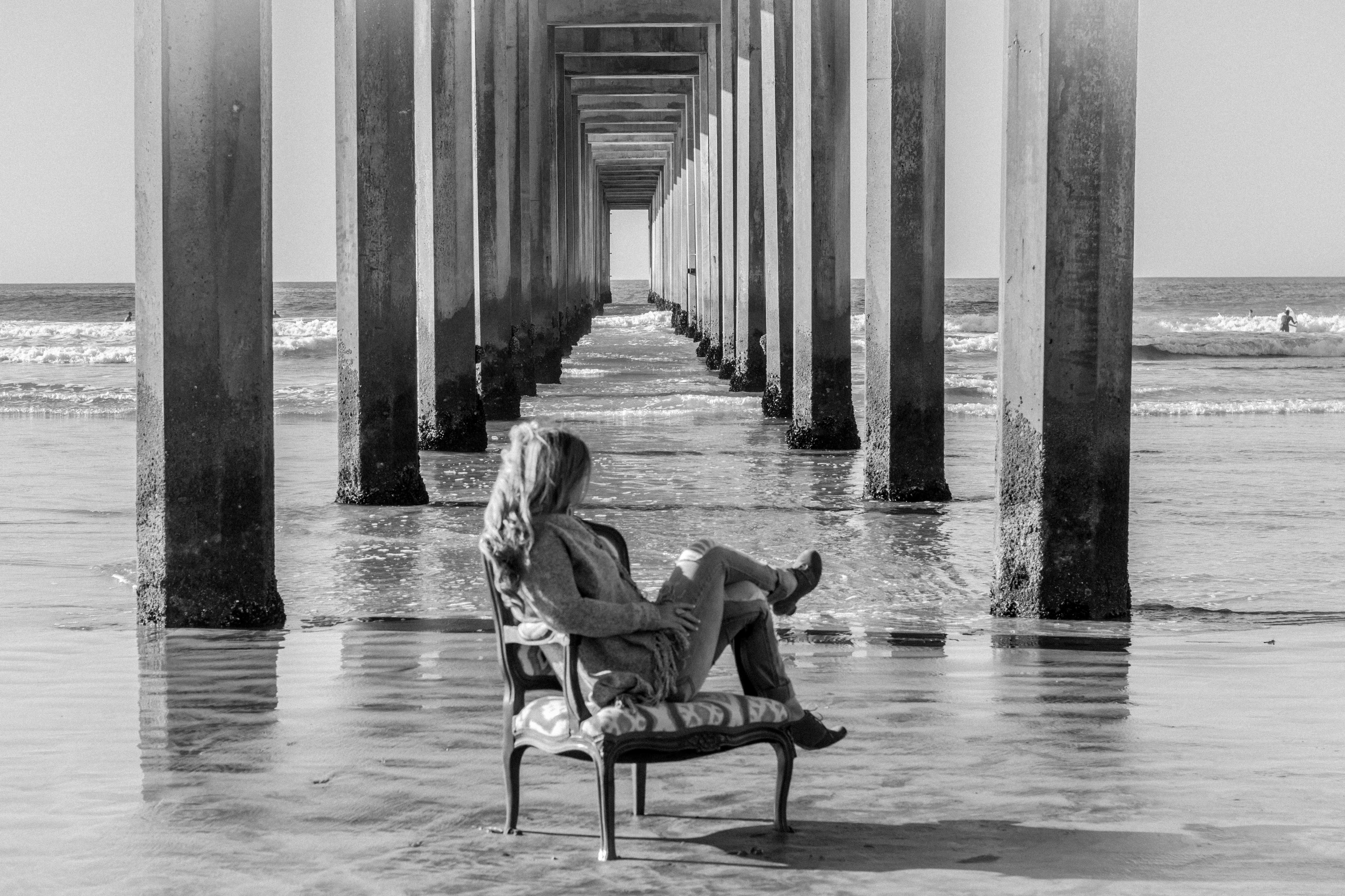 Black-and-white photo of a woman relaxing in an elegant French-style upholstered chair on the sand beneath a long pier, gazing at the ocean